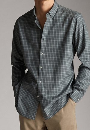 Homme portant une chemise à carreaux grise à manches longues et un pantalon beige, les mains dans les poches, sur un fond neutre.
