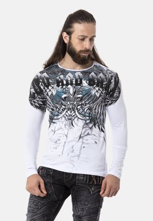 Langärmliges weißes Hemd mit detailliertem schwarz-blauen Grafikdesign, das Flügel und ein Fleur-de-Lis beinhaltet, mit einem bedruckten abgenutzten Effekt.
