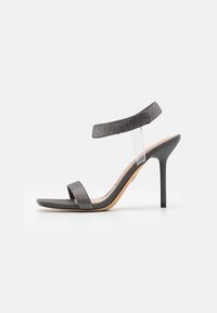 Musta korkokenkä, jossa on läpinäkyvä hihna, teksturoitu päällinen ja kapea stiletto-korko. Ominaisuuksiin kuuluu leveä etuhihna ja kiiltävät yksityiskohdat.