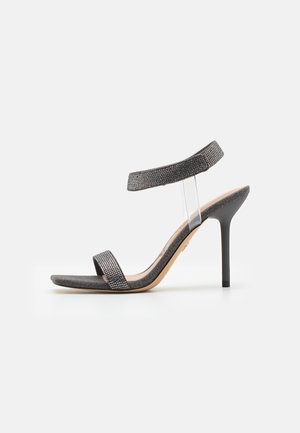 Black heeled sandal with a transparent strap, textured upper, and slim stiletto heel. Features a wide front band and shimmery accents.