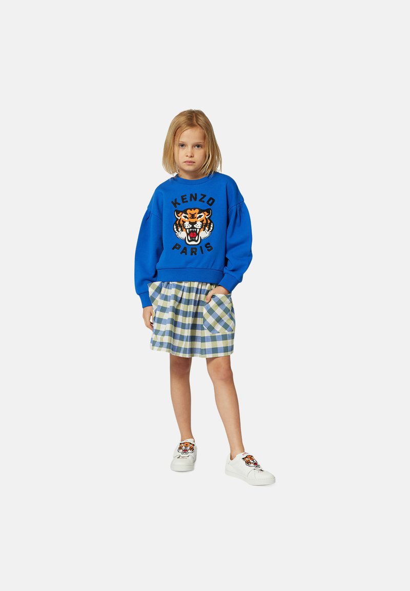 Sweatshirt bleu avec graphique de tigre, associé à une jupe écossaise dans des tons verts et bleus ; baskets blanches avec détail de tigre.
