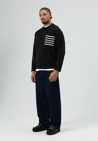 Zwarte trui met lange mouwen en een borstzak met witte strepen, gecombineerd met losse donkeren denim broek en zwarte laarzen met roze veters. Simpele achtergrond.