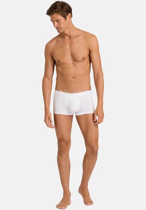 Erwachsener Mann steht barfuß in schlichten weißen Boxer-Shorts und blickt nach unten links vor einem hellgrauen Hintergrund.