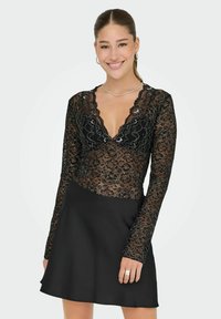 Svart spetsbodysuit med långa ärmar och scalloped halsringning, parat med en svart A-linje kjol. Spetsen har blommönster och texturerad detalj.