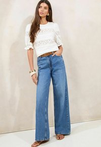Blusa bianca in pizzo con maniche a sbuffo, abbinata a jeans in denim blu a vita alta e gamba larga. Una cintura marrone e sandali piatti completano il look.