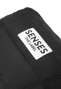 Schwarzer, gesteppter Steppstoff mit einem weißen Etikett, das "SENSES" und "THE LABEL" in schwarzem Text zeigt, und hebt das Logodesign sowie die Textur hervor.