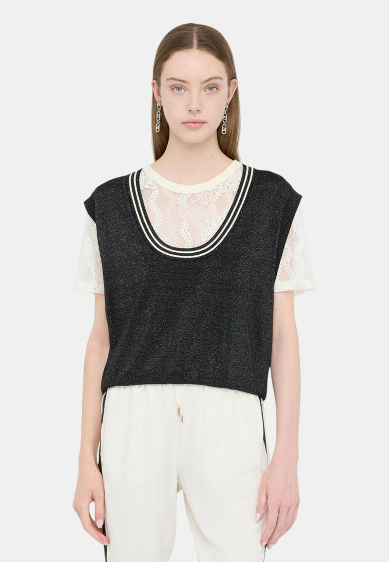 Top nero senza maniche in tessuto knit, indossato sopra una camicia bianca in pizzo. Presenta un ampio scollo rotondo con strisce nere a contrasto.