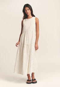 Robe maxi sans manches, crème, à pois noirs, taille cintrée et jupe à volants. Portée avec des sandales plates noires. Texture lisse.