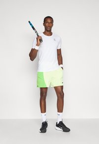 Λευκό αθλητικό μπλουζάκι κοντό μανίκι, ανοιχτοπράσινο σορτσάκι με μαύρες λεπτομέρειες, μαύρα αθλητικά παπούτσια, κρατώντας μια ρακέτα τένις, με βραχιόλια στους δύο καρπούς.