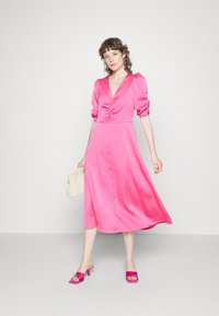 Vestido midi de cetim cor-de-rosa brilhante com mangas bufantes, decote em V e detalhe franzido na parte do busto. A modelo usa sandálias cor-de-rosa de slide e segura uma bolsa texturizada.