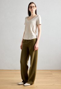 Tricou bej cu decolteu în formă de V și mâneci scurte, purtat cu pantaloni largi de culoare verde măsliniu. Pantofi stilați albi completează ținuta. Podea din lemn natural.