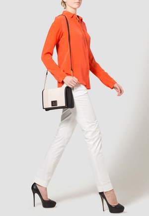 Femme marchant, vêtue d'un chemisier orange à manches longues, d'un pantalon blanc, de talons hauts noirs, et portant un sac à épaule noir et blanc.