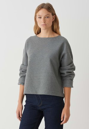 Grau, lässiger Pullover mit rundem Halsausschnitt, langen Ärmeln und umgeschlagenen Bündchen, kombiniert mit dunklen Jeans. Die weiche Stofftextur ist sichtbar.
