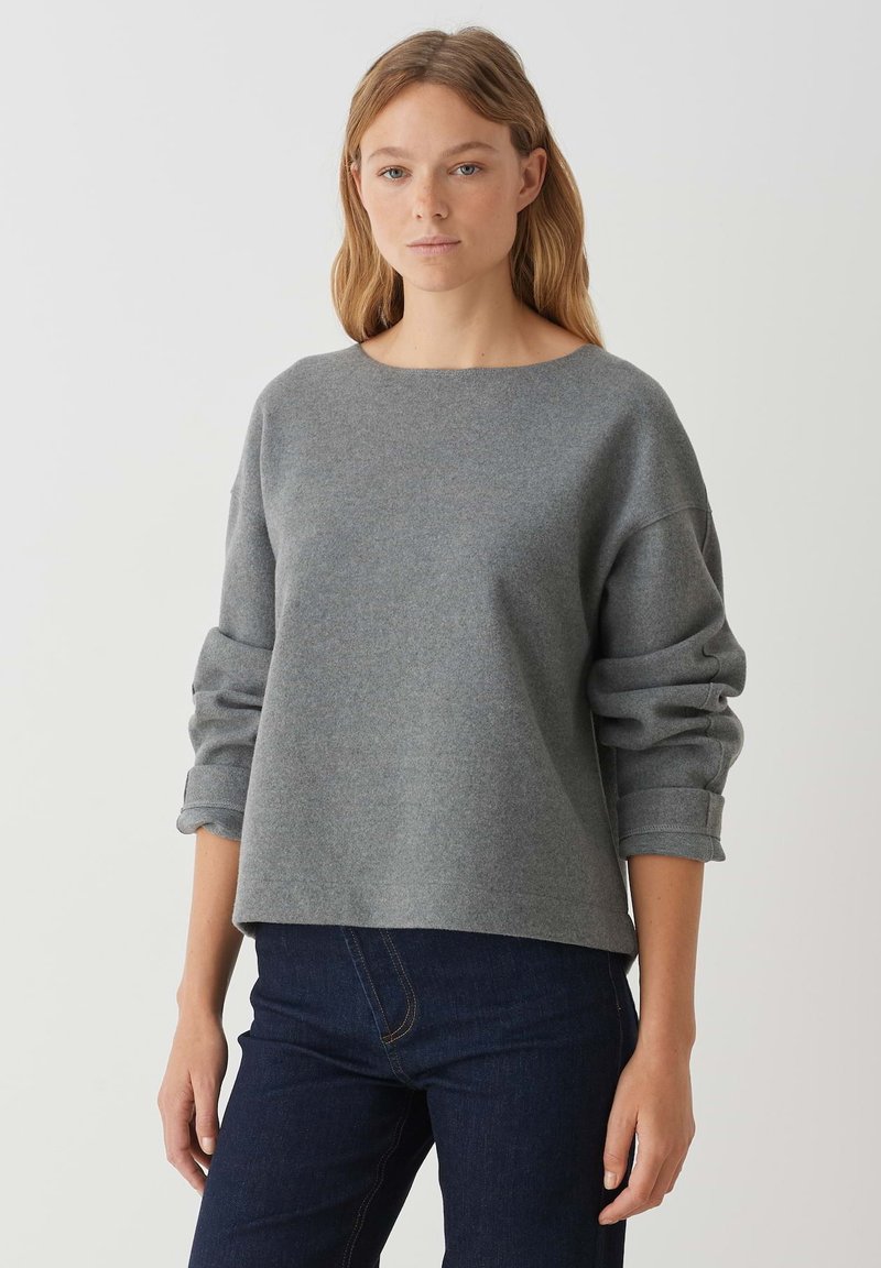 Grau, lässiger Pullover mit rundem Halsausschnitt, langen Ärmeln und umgeschlagenen Bündchen, kombiniert mit dunklen Jeans. Die weiche Stofftextur ist sichtbar.