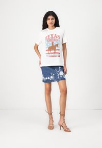 Hvit t-skjorte med "Texas Rodeo" grafikk i rød, blå og brun. Denim skjørt med blomsterbroderi i hvitt, paired med nude stroppesandaler.