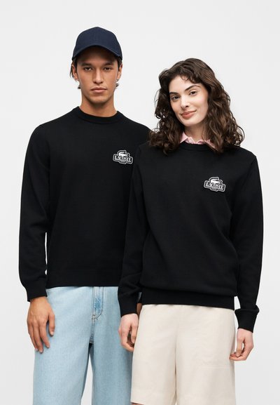 Homme et femme portant des pulls noirs Lacoste, l'homme avec une casquette bleue et un jean, la femme avec un col de chemise rose visible et un short beige, debout côte à côte.