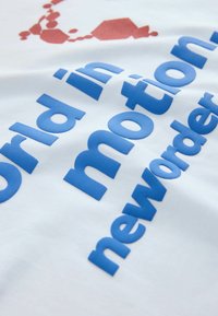 Texte sur tissu blanc affichant « monde en mouvement » en lettres bleues épaisses et « nouvel ordre » en lettres bleues plus petites, avec une forme abstraite rouge au-dessus.