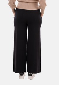 Persona che indossa pantaloni larghi neri, un maglione beige e sneaker bianche, in piedi con le mani nelle tasche e con la schiena rivolta.
