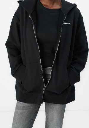 Person, der einen schwarzen Kapuzenpullover mit Reißverschluss trägt, Hände in den Taschen, schwarzes Oberteil darunter und graue Jeans vor einem schlichten weißen Hintergrund.