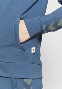 Niebieski sweatshirt z boczną kieszenią, teksturowanym zielonym wzorem w chevron na rękawie oraz małą metką u dołu. Miękka tkanina, nowoczesny krój.