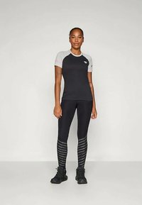 Kobieta stoi, ubrana w czarną i szaro-skalną koszulkę sportową z krótkim rękawem, czarne legginsy z szarymi paskami przy łydkach oraz czarne buty sportowe.