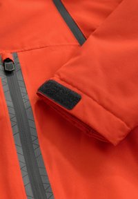 Giacca outdoor rossa con una trama liscia, zip nere e dettagli in velcro sui polsini. Include motivi geometrici sui cursori delle zip.