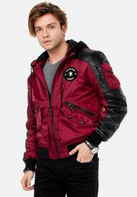 Rote Satin-Bomberjacke mit schwarzen Leder-Schultern, mehreren Reißverschlusstaschen, Kordelzügen an der Kapuze und einem runden Logo auf der Brust.