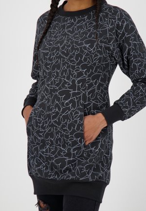 Frau trägt ein schwarzes langes Sweatshirtkleid mit hellgrauem abstraktem Herzlinienmuster, die Hände in den Vorderentaschen und schwarzen gerippten Bündchen sowie Saum.