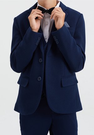 Persona ajustando una pajarita negra, vistiendo un traje azul marino texturizado con camisa blanca y un pañuelo de bolsillo estampado sobre fondo blanco.