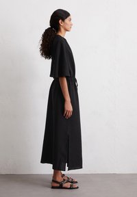 Abito nero lungo fino alle caviglie con maniche corte e larghe, vita cinchata e spacchi laterali. La modella indossa sandali neri con cinturino. Tessuto semplice e texture.