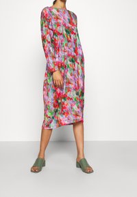 Blumen-Midi-Kleid mit langen Ärmeln, das leuchtend rote, grüne und lila Aquarellmuster aufweist. Kombiniert mit grünen offenen Blockabsatzschuhen.