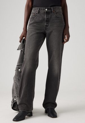 Persoon met een high-waisted wijd uitlopende broek van vervaald zwart denim, zwarte laarzen met hak en vierkante neus, die een bijpassend vervaald zwart denim jack vasthoudt.