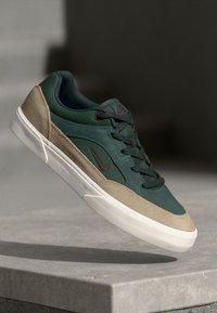 Sneaker featuring a green leather upper, tan suede accents, black laces, and a white rubber sole, with a small burgundy logo on the side.