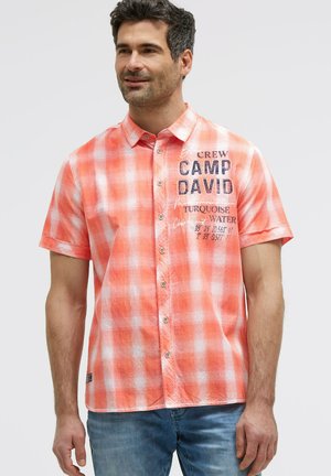 Mann trägt ein kurzärmliges orange-weiß kariertes Hemd mit Text auf der Brust und blaue Jeans.