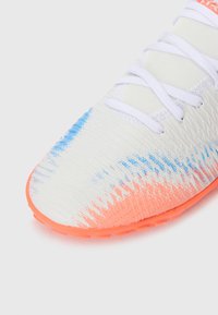 Bout d'une chaussure de sport blanche avec une semelle orange et des détails de peinture en dégradé bleu et orange, lacets blancs sur un fond uni.
