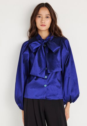 Mėlyna satino bluzelė su dideliu raiščiu prie apykaklės, pūstuosiais rankovėmis ir dekoratyviniais mygtukų akcentais. Lygus paviršius su blizgiu apdailos efektu.