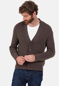 Cardigan marron côtelé avec un col en V, doté de cinq boutons marron, manches longues et une finition texturée. Porté sur une chemise blanche.