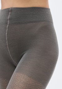 Graue Leggings mit einem gerippten hohen Bund und einer glatten Textur, die in einen transparenten, gemusterten Beinbereich am unteren Ende übergehen.