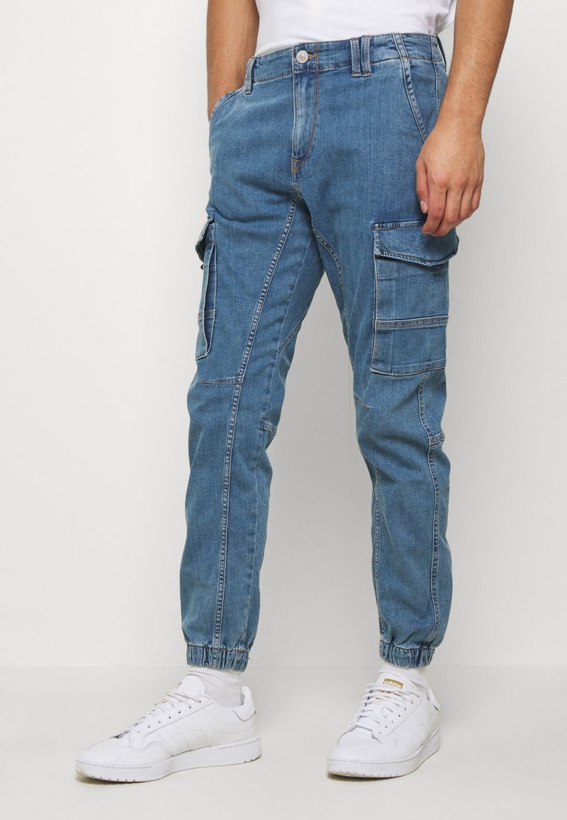 Pantalones cargo de mezclilla azul claro con textura ripstop, puños elásticos y múltiples bolsillos laterales, combinados con zapatillas blancas.