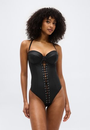 Ženska kodrastih las, oblečena v črn, spredaj zavezani, brez naramnic korzet bodi na enostavni belem ozadju.