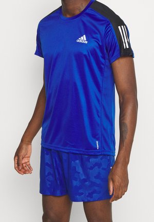Mann trägt blaues Adidas-Sportshirt mit schwarzen Ärmeln und blau gemusterten Shorts, steht vor einfachem Hintergrund.