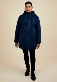 Parka isolata blu navy con cappuccio, vita regolabile con cordino e zip frontale. Abbinata a pantaloni neri e stivali alla caviglia, su uno sfondo chiaro.