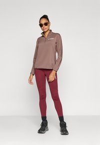 Barna hosszú ujjú pulóver gallérral és logóval, bordó leggingsszel kontrasztos varrásokkal, fekete sportcipővel párosítva.