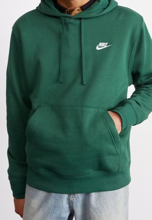 Mężczyzna w zielonej bluzie z kapturem Nike z kieszenią typu kangur i sznurkami, w połączeniu z jasnoniebieskimi dżinsami, ręka w kieszeni.