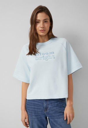 Giovane donna con capelli castani lisci indossa una felpa azzurra oversize a maniche corte con la scritta "bloom bright" e jeans blu.