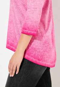 Die Hand einer Frau ruht auf dem Oberschenkel einer schwarzen Jeans und trägt ein pinkfarbenes Oberteil mit drei Viertelärmeln und einem dunkelpinkfarbigen Saum.