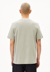 Person mit glatze, der ein hellgraues kurzärmliges Hemd und dunkle Hosen trägt, von hinten gezeigt, wobei ein Tattoo auf dem rechten Arm sichtbar ist.