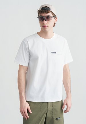 Młody mężczyzna w białej koszulce Columbia, oliwkowo-zielonych spodniach, beżowej czapce z daszkiem noszonej do tyłu oraz ciemnych sportowych okularach przeciwsłonecznych.