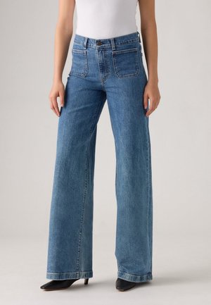 Kvinde iført højtaljede, vide blå jeans med forlommer, kombineret med sorte spidse hæle og en hvid ærmeløs top.