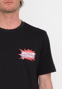 Svart kortärmad t-shirt med en färgglad "VOLCOM & The STONES" grafik på vänster bröst, med en stjärnklara bakgrund.